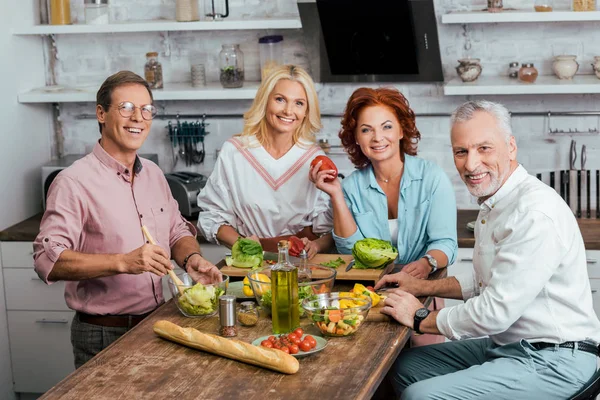 Sonrientes amigos maduros preparando ensalada para la cena en casa - foto de stock