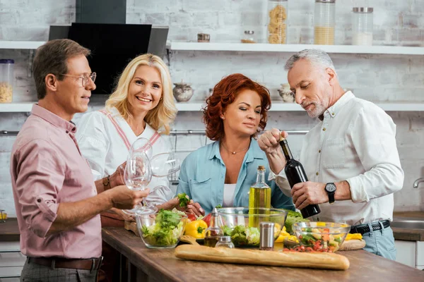 Souriant amis matures célébrer la réunion avec la salade et le vin dans la cuisine — Photo de stock