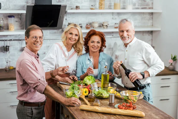 Ältere Freunde feiern Treffen mit Salat und Wein in der Küche, blicken in die Kamera — Stockfoto