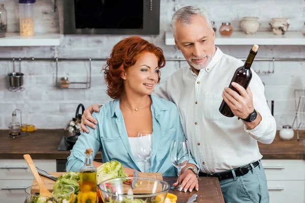 Husband hugging wife during salad preparation for dinner in kitchen and holding wine bottle — Stock Photo