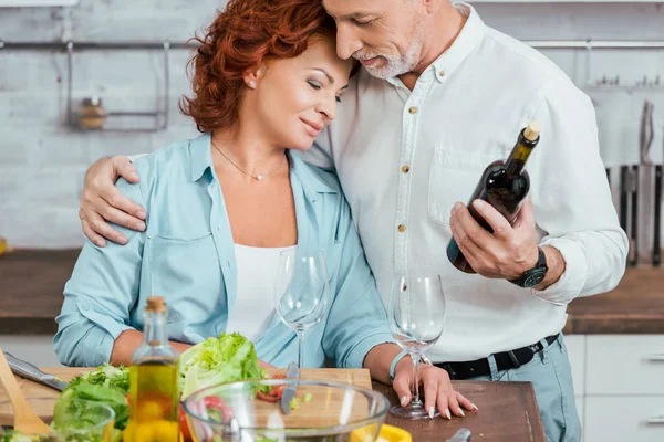 Marido afetuoso abraçando esposa durante a preparação da salada para o jantar na cozinha e segurando garrafa de vinho — Fotografia de Stock