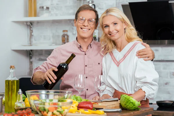 Smiling husband hugging wife and pouring wine during dinner at home, looking at camera — Stock Photo