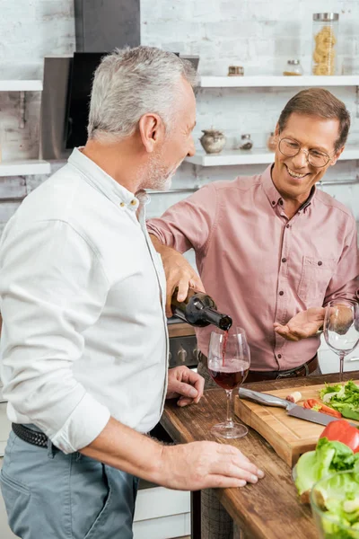 Schöner glücklicher Mann schenkt einem alten Freund beim Abendessen in der Küche Wein ein — Stockfoto