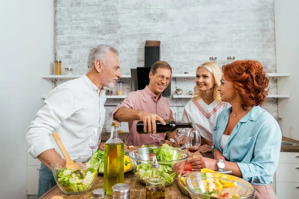 Schöner lächelnder Mann schenkt glücklichen alten Freunden beim Abendessen in der Küche Wein ein — Stockfoto