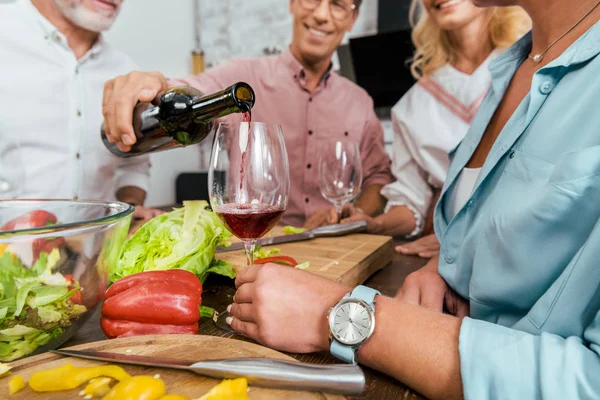 Imagen recortada de hombre guapo vertiendo vino a viejos amigos felices durante la cena en la cocina - foto de stock