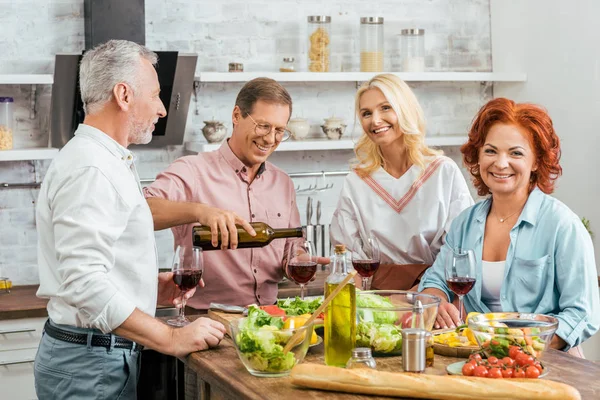 Schöner Mann schenkt glücklichen alten Freunden beim Abendessen in der Küche Wein ein — Stockfoto