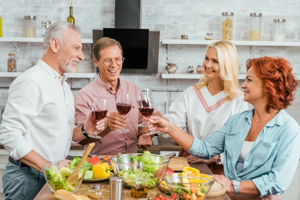 Viejos amigos sonrientes tintineo con gafas de vino en la cocina - foto de stock