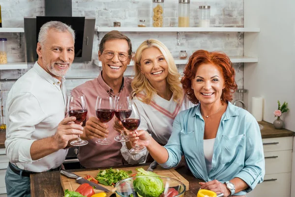 Sonriendo viejos amigos tintineo con gafas de vino en la cocina y mirando a la cámara - foto de stock