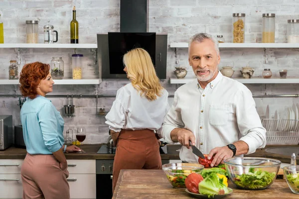 Handsome man preparing salad for dinner at home, women talking with wine — Stock Photo