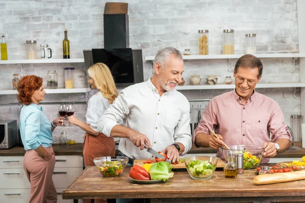 Beaux hommes souriants préparant la salade pour le dîner à la maison, femmes parlant avec du vin — Photo de stock