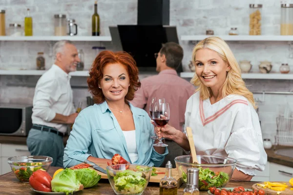 Smiling beautiful women preparing salad for dinner, holding wineglasses and looking at camera at home — Stock Photo