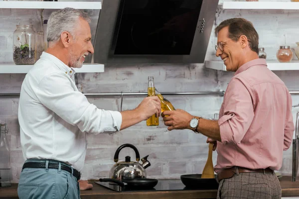 Side view of smiling handsome men clinking with bottles of beer while cooking at home — Stock Photo