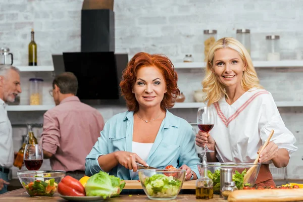 Lächelnde schöne Frauen, die Salat für das Abendessen mit Wein zubereiten und zu Hause in die Kamera schauen — Stockfoto