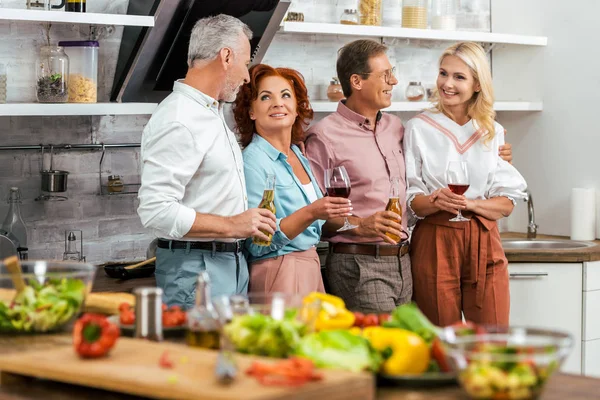 Vecchi amici che tengono bevande alcoliche e parlano in cucina, verdure sul tavolo — Foto stock