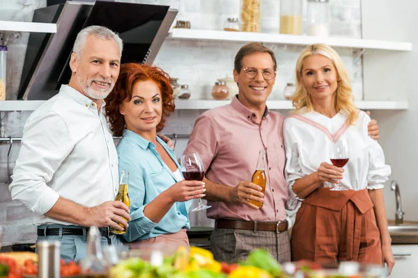 Happy mature friends holding glasses of wine and beer bottles, smiling at camera in kitchen — Stock Photo
