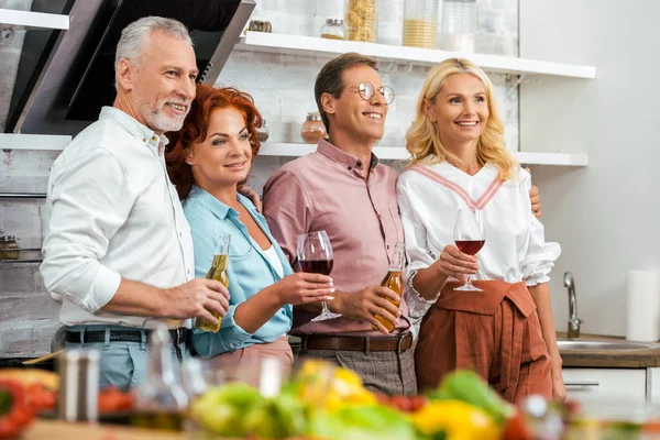 Felizes amigos maduros segurando bebidas alcoólicas e olhando para longe na cozinha — Fotografia de Stock