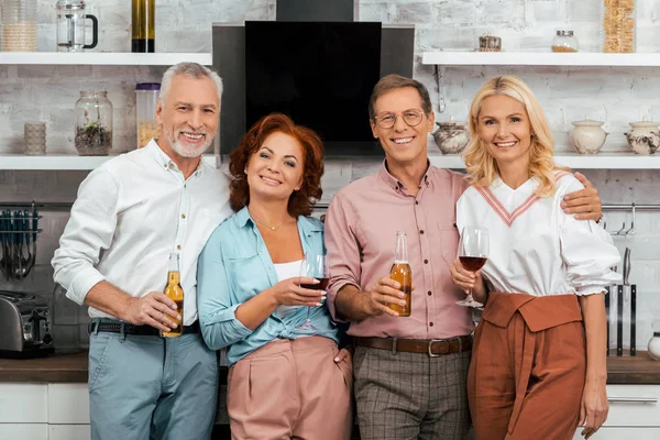 Felices amigos maduros sosteniendo bebidas alcohólicas y sonriendo a la cámara en la cocina - foto de stock