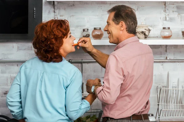 Back view de homem sorridente alimentando bela mulher madura enquanto estão juntos na cozinha — Fotografia de Stock