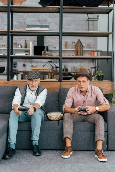 Mature male friends using joysticks and looking at camera while sitting together on couch — Stock Photo