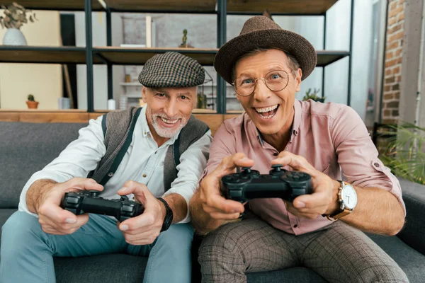 Hombres maduros emocionados jugando con joysticks y sonriendo a la cámara - foto de stock