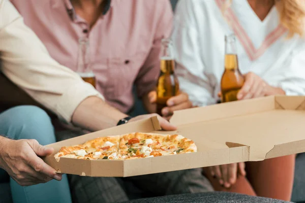 Recortado disparo de hombre sosteniendo caja de pizza y amigos bebiendo cerveza detrás - foto de stock
