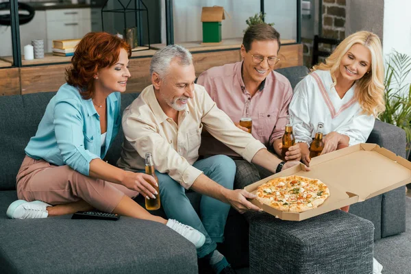 Sorridenti amici maturi che bevono birra e mangiano pizza seduti sul divano di casa — Foto stock