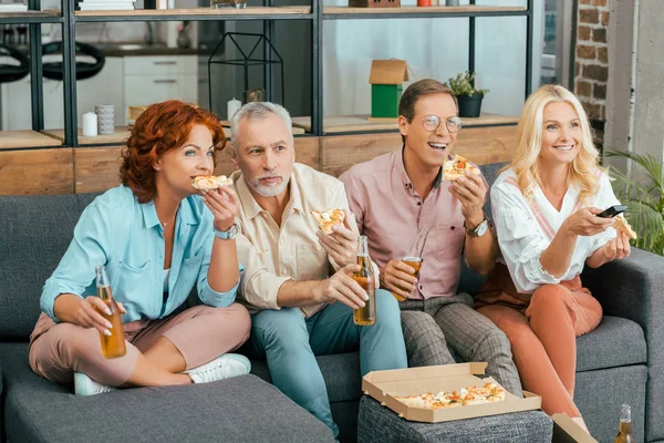 Happy mature friends eating pizza and drinking beer while using remote controller and watching tv together at home — Stock Photo