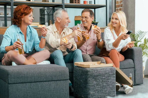 Happy mature men and women eating pizza and drinking beer while using remote controller and watching tv together at home — Stock Photo