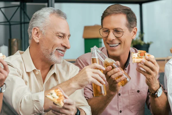 Feliz maduro los hombres comer pizza y tintineo cerveza botellas en casa - foto de stock