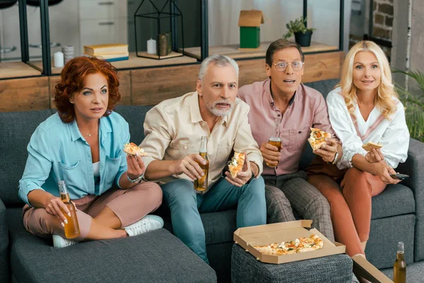 High angle view of mature friends sitting on sofa, drinking beer and eating pizza while watching tv at home — Stock Photo