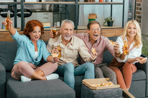 Amigos maduros emocionados bebiendo cerveza y comiendo pizza en casa - foto de stock