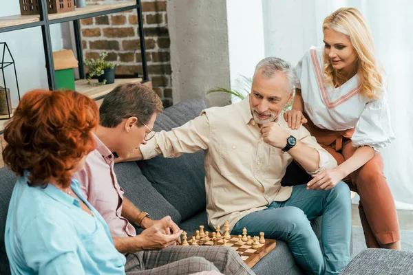 Дві зрілі пари сидять на дивані і грають в шахи разом вдома — стокове фото