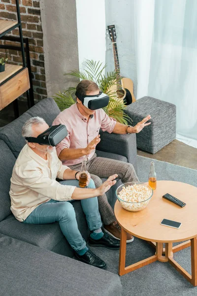 Visão de alto ângulo de homens maduros bebendo cerveja e usando fones de ouvido de realidade virtual enquanto sentados no sofá — Fotografia de Stock