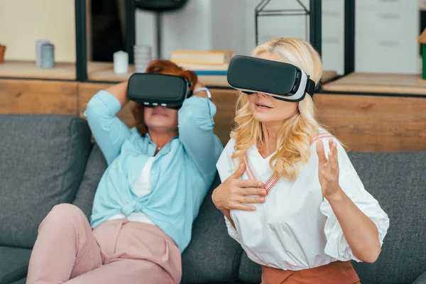 Hermosas mujeres maduras sentadas en el sofá y el uso de auriculares de realidad virtual - foto de stock