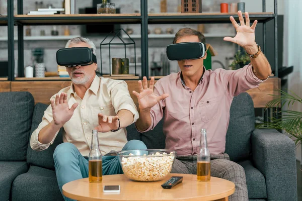 Amigos masculinos maduros usando auriculares de realidad virtual mientras beben cerveza y comen palomitas de maíz en casa - foto de stock
