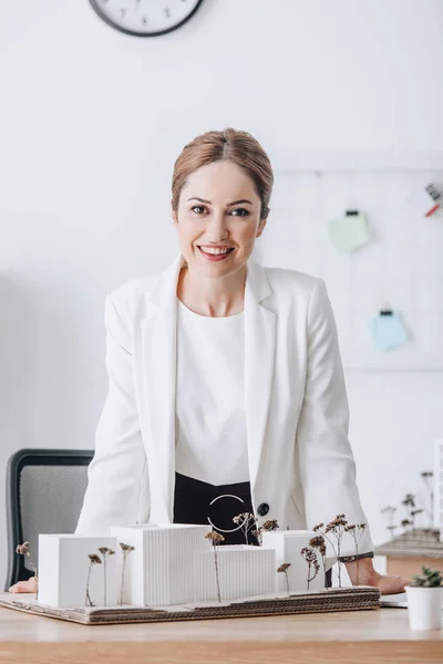 Cheerful female architect with house model looking at camera at workplace — Stock Photo