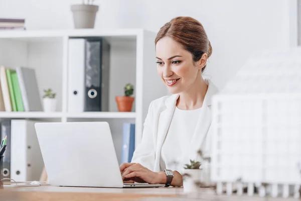 Sorridente arquiteto feminino trabalhando com laptop no local de trabalho com modelo de casa, foco seletivo — Fotografia de Stock