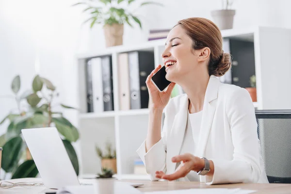 Laughing businesswoman talking on smartphone in office with laptop — Stock Photo