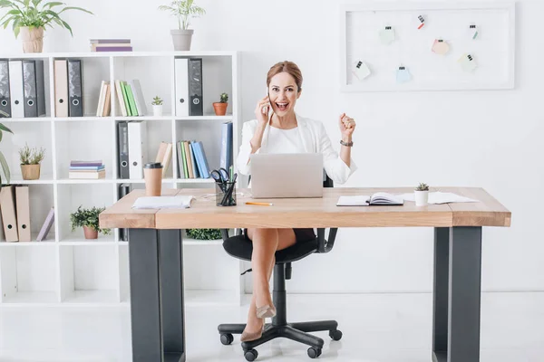 Successful professional business woman talking on smartphone while triumphing in modern office with laptop — Stock Photo