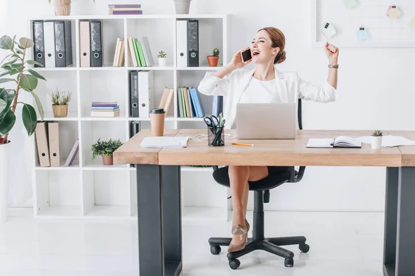Успішна бізнес-леді розмовляє на смартфоні під час тріумфу в сучасному офісі з ноутбуком — стокове фото