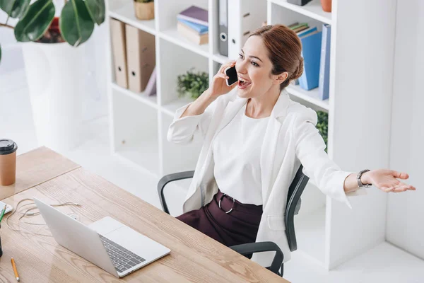 Aufgeregte Geschäftsfrau spricht im modernen Büro mit Laptop auf Smartphone — Stockfoto