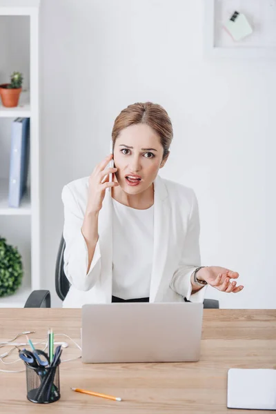 Confused emotional businesswoman talking on smartphone at workplace with laptop — Stock Photo