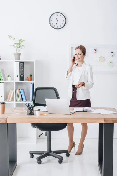 Exitosa mujer de negocios hablando en el teléfono inteligente en la oficina moderna con el ordenador portátil - foto de stock