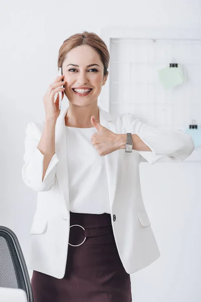 Alegre mujer de negocios hablando en smartphone y mostrando el pulgar hacia arriba en la oficina - foto de stock