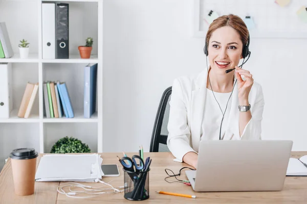 Operador feminino profissional atraente com fone de ouvido e laptop no escritório moderno — Fotografia de Stock