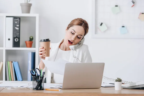 Schockierte Managerin hält Coffee to go, während sie telefoniert und im Büro mit Laptop arbeitet — Stockfoto