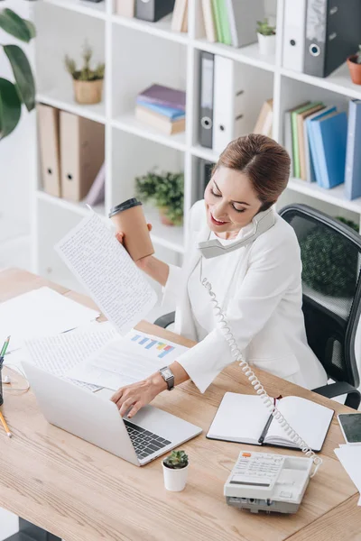 Lächelnde Geschäftsfrau mit Dokumenten und Coffee to go, während sie telefoniert und im Büro mit Laptop arbeitet — Stockfoto