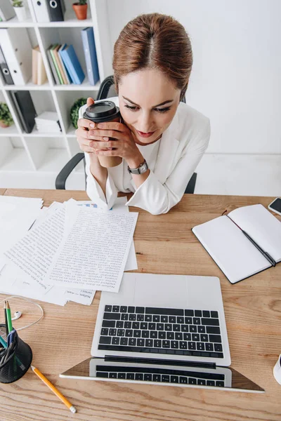 Vista aérea de la mujer de negocios con café para ir a trabajar con los documentos y el ordenador portátil mientras está sentado en la oficina moderna - foto de stock