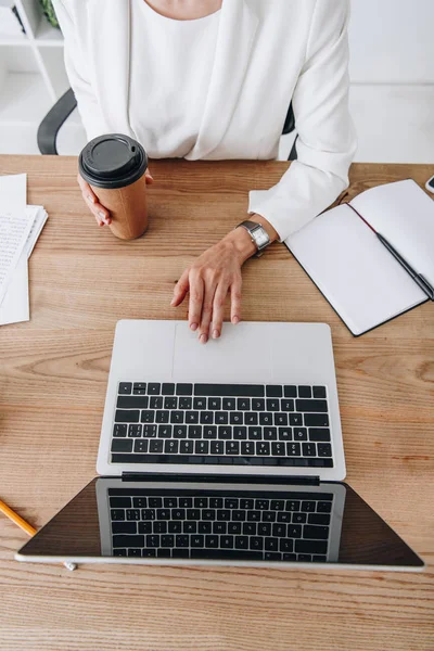 Vue supérieure recadrée de femme d'affaires avec café pour aller travailler avec un ordinateur portable et des documents sur le lieu de travail — Photo de stock