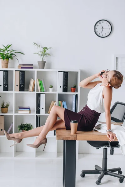 Zufriedene Geschäftsfrau, die während der Kaffeepause im modernen Büro auf dem Tisch sitzt und mit dem Smartphone spricht — Stockfoto